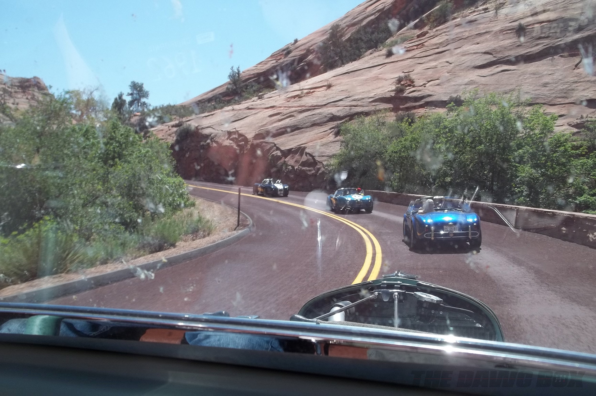 one black and 2 blue Shelby AC Cobras driving down a scenic Grand Canyon highway