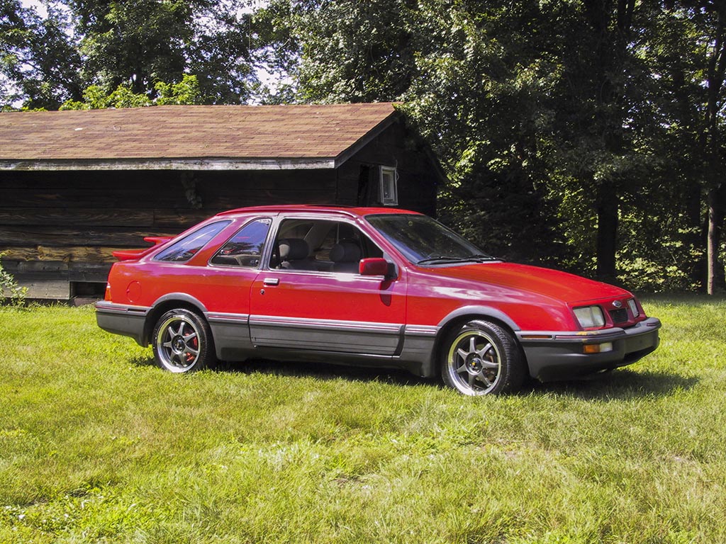Red ford Merkur XR4Ti with custom wheels parked next to a cabin